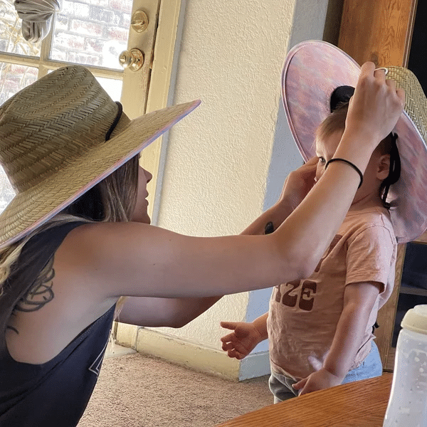 Mother helping toddler wear a large straw hat with pink trim, showcasing custom hats for family matching, ideal for outdoor activities and sun protection.