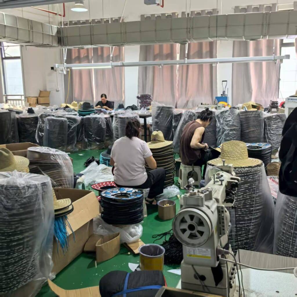 Factory workers assembling and stitching custom straw hats, surrounded by stacks of wide brim lifeguard hats ready for packaging.