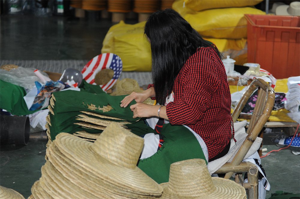 Craftsman assembling straw hats in a workshop, with stacks of unfinished hats and various materials in the background.